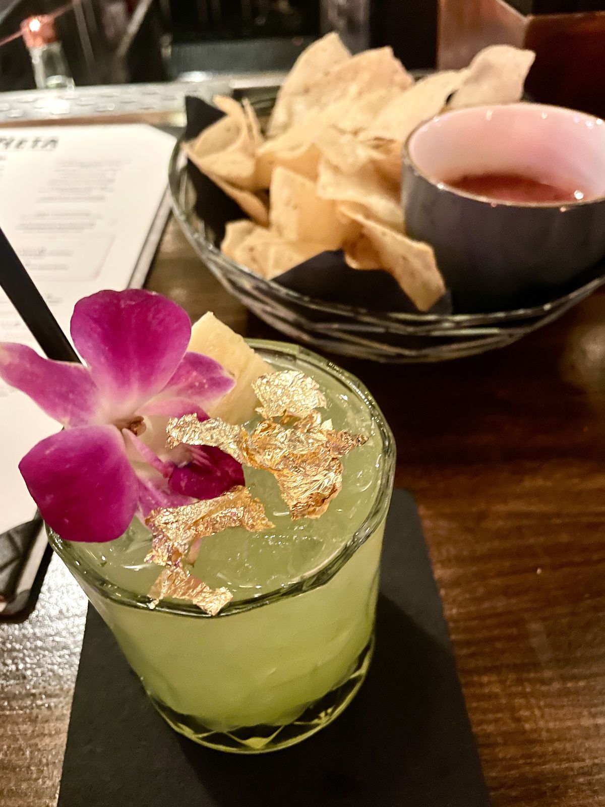 A bartop holds a puce cocktail with a flower and gold foil. Next to it on the right back are chips and salsa. A menu can be seen to the left.