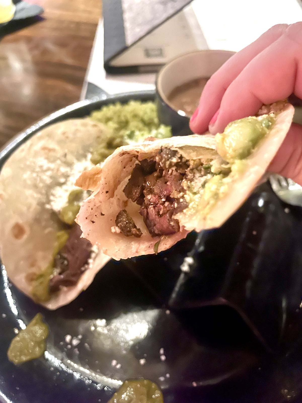 A steak taco with a bite taken out and lots of cheese and guacamole is held by a woman. A second taco sits on a plate.