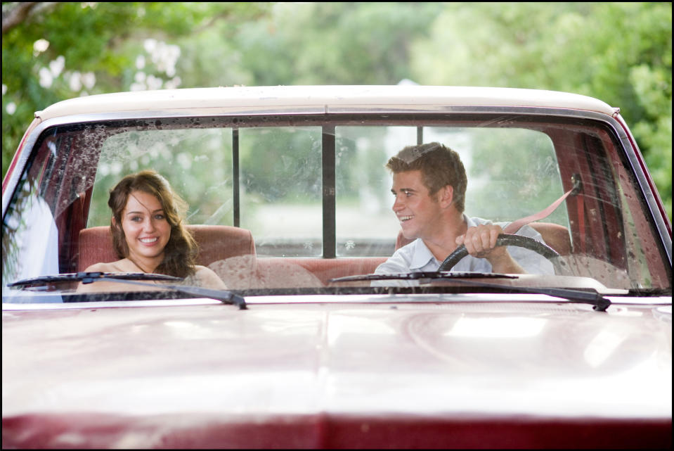 Miley Cyrus and Liam Hemsworth sit in an old truck. Miley smiles, seated on the passenger side, while Liam is at the wheel, looking at her and smiling