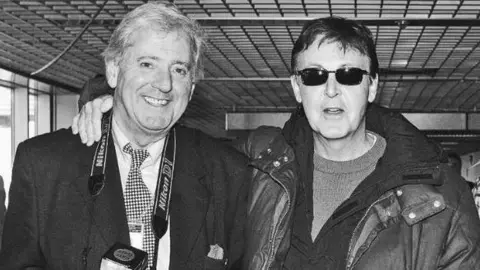 Dennis Stone Paul McCartney wearing a jacket and sunglasses has an arm around Dennis Stone, who wears a camera around his neck, at Heathrow Airport